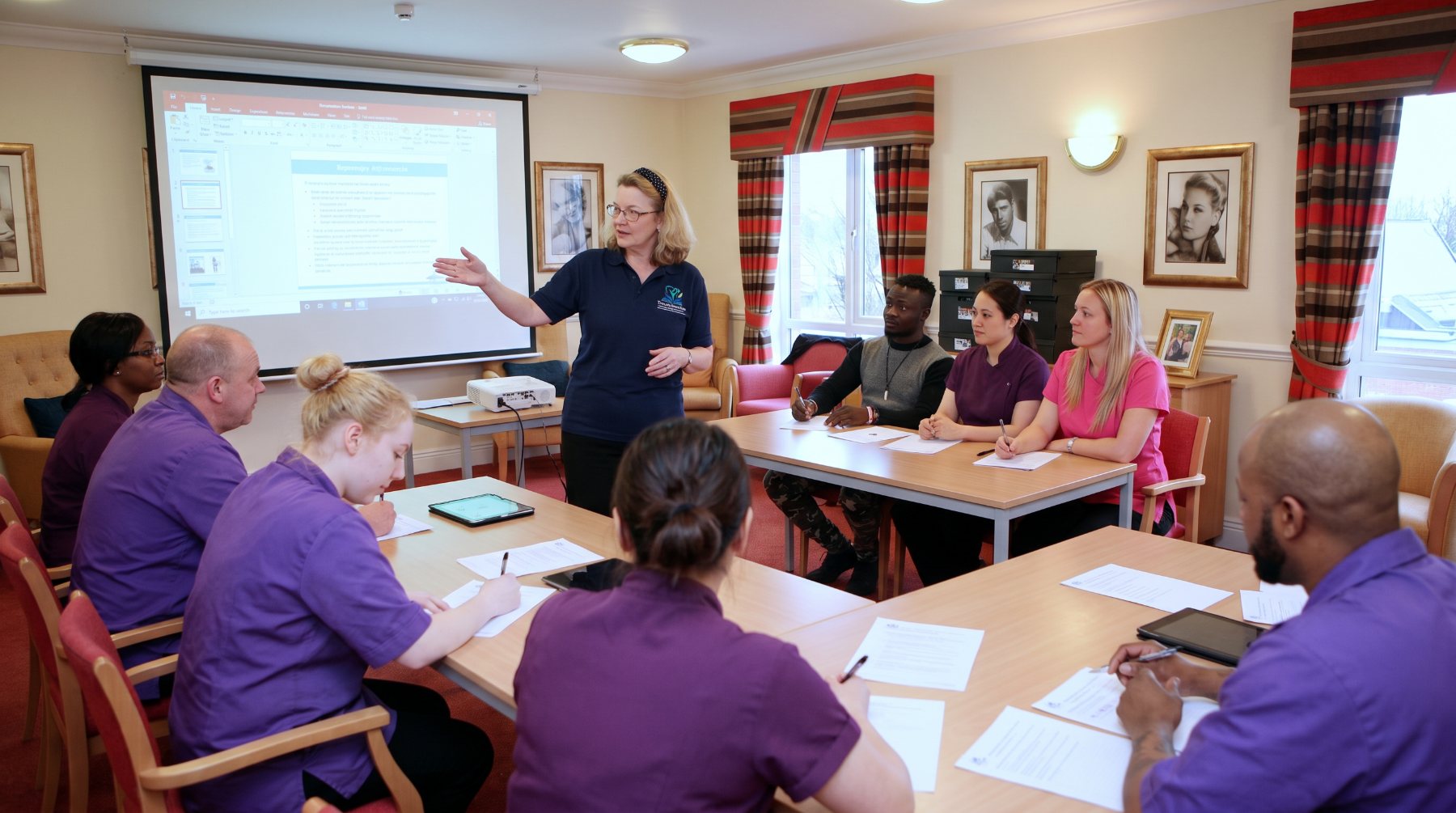 A TransformCare trainer leading a session for a culturally diverse group of care staff seated around tables in a care home lounge