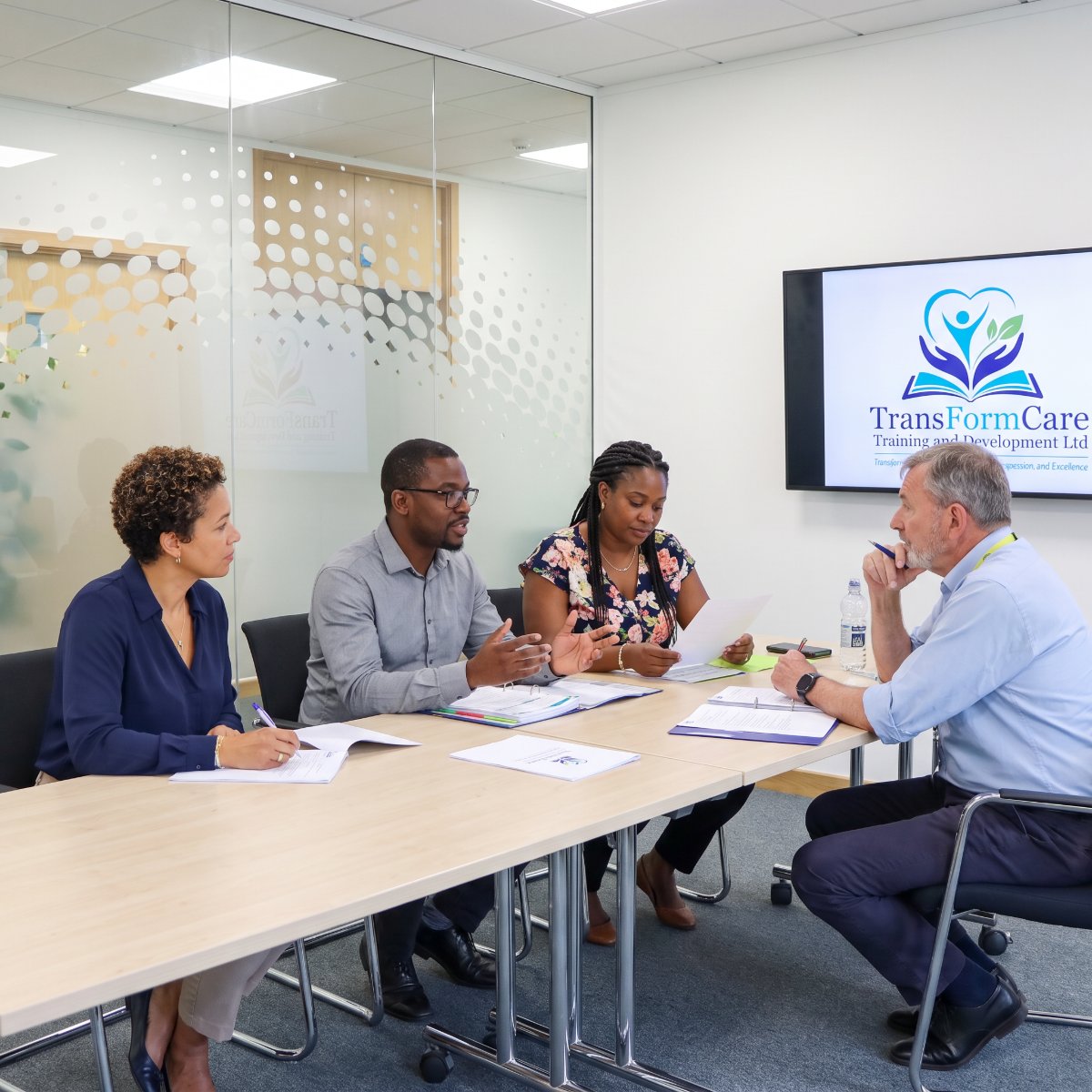 Care managers around a meeting table reviewing training documents, with a TransformCare slide on the screen behind them